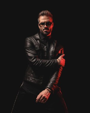 Güneş gözlüklü seksi sakallı adam siyah arka planda dururken kırmızı ışıklarla havalı poz veriyor.