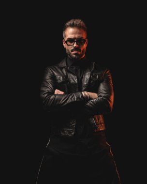 Siyah deri ceketli dramatik havalı adam kollarını kavuşturup siyah arka planda ileriye bakıyor.