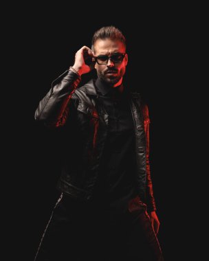 Siyah deri ceketli gizemli adam güneş gözlüğü takıyor ve siyah arka planda duruyor.