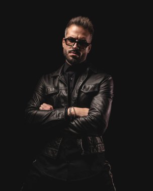 Güneş gözlüklü kendine güvenen dramatik bir adam kollarını kavuşturup siyah arka planda ileriye bakıyor.