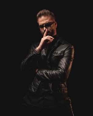 Güneş gözlüklü yakışıklı adam siyah arka planda dururken sessiz hareketler yapıyor ve kollarını kavuşturuyor.