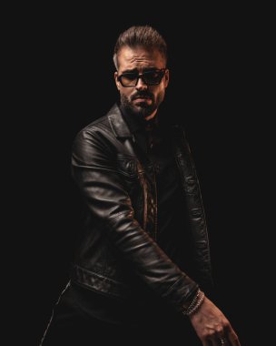 Siyah ceketli, güneş gözlüklü, siyah arka planda poz veren havalı, dramatik bir adam.