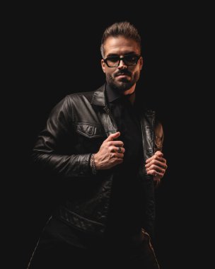 Güneş gözlüklü seksi adam siyah arka planda dikilirken siyah deri ceketi tamir ediyor.