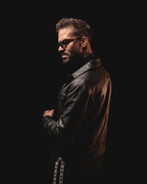 Siyah deri ceketli, güneş gözlüklü, siyah arka planda duran seksi sakallı adamın yan görüntüsü.