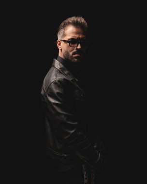 Siyah deri ceketli ve siyah arka planda duran havalı moda adamının yan görüntüsü.