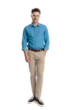 Mavi gömlekli ve gözlüklü mutlu genç adam, elleri ceplerinde duruyor ve gülümsüyor, beyaz arka planda soyutlanmış..