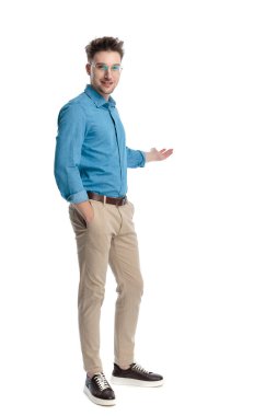 Mavi gömlekli ve gözlüklü mutlu genç adam, bir eli cebinde duruyor ve beyaz arka planda izole edilmiş bir şekilde sunum yapıyor..