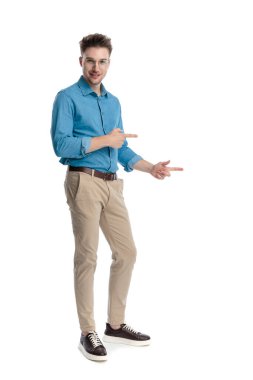 Mavi tişörtlü ve gözlüklü mutlu genç adam, iki eli yanda duruyor ve işaret ediyor, beyaz arka planda izole edilmiş..