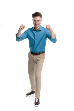 Mavi gömlekli ve gözlüklü mutlu genç adam, ayakta duruyor ve kutluyor, iki eli havada, beyaz arka planda izole edilmiş..