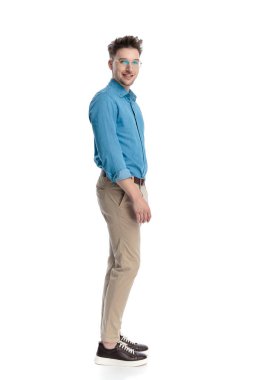 Gözlüklü, ayakta duran ve beyaz arka planda izole edilmiş kameraya bakan mutlu genç bir adamın yan görüntüsü..