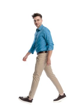 Gözlüklü, gülümseyen ve yürüyen, beyaz arka planda izole bir şekilde kameraya bakan mutlu genç bir adamın yan görüntüsü..