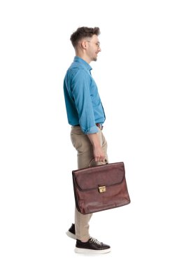 Gözlüklü, çantalı, ayakta duran, gülümseyen, beyaz arka planda soyutlanmış mutlu genç bir adamın yan görüntüsü..