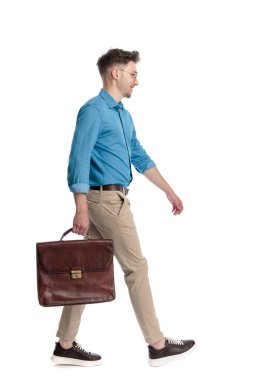 Gözlüklü, elinde evrak çantası olan ve beyaz arka planda izole bir şekilde yürüyen mutlu genç bir adamın yan görüntüsü..