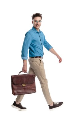 Gözlüklü ve evrak çantalı mutlu genç bir adam, beyaz arka planda izole bir şekilde yürüyordu ve arkasını kolluyordu..