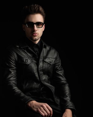 Siyah deri ceketli, güneş gözlüklü esrarengiz adam siyah arka planda oturuyor.