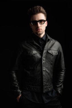 Güneş gözlüklü çekici bir genç adam ceplerinde el ele tutuşuyor ve siyah arka planda ileriye bakıyor.