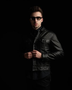 Seksi genç adam siyah deri ceketi güneş gözlükleriyle ayarlarken siyah arka planda dikilip ileriye bakıyor.