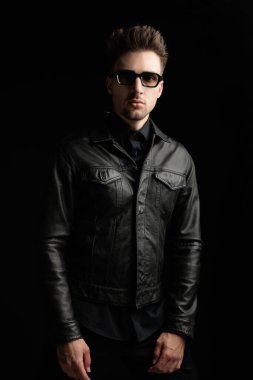 Siyah deri ceketli, siyah gözlüklü gizemli genç adam ileri bakıyor ve siyah arkaplanın önünde duruyor.
