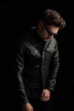 Siyah deri ceketli, güneş gözlüklü, siyah arka planda poz veren havalı bir adam.