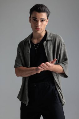 Gri hırkalı yakışıklı esmer adamın portresi avuçlarını ovuyor ve gri arka planda ileriye bakıyor.