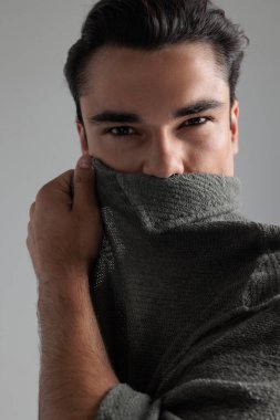 Gri arkaplan önünde dururken ağzı gri hırkayla kaplayan genç adamın portresini kes.