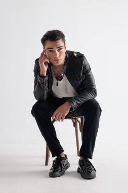 Siyah deri ceketli seksi genç adam tapınağa dokunuyor, ileri bakıyor ve gri arka planda oturuyor.