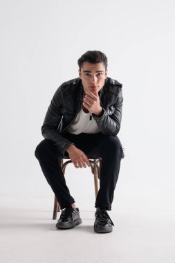 Siyah deri ceketli düşünceli genç adam çenesine dokunuyor, düşünüyor ve beyaz arka planda oturuyor.