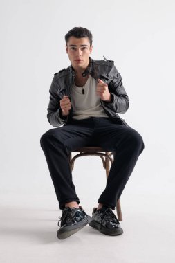 Çekici genç adam ileriye bakıyor, siyah deri ceketini ayarlıyor ve gri arka planda oturuyor.