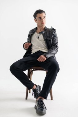 Siyah deri ceketli yakışıklı havalı adam gri arka planda oturuyor. 