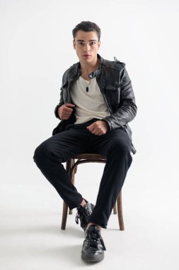 Siyah deri ceketli genç yakışıklı adam ileri bakıyor ve gri arka planda oturuyor.