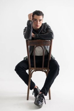 Siyah deri ceketli seksi, kendine güvenen adam koltukta kollarını tutuyor ve gri arka planda otururken ileriye bakıyor.