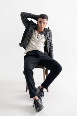 Siyah deri ceketli seksi genç adam oturuyor ve elleri başının arkasında gri arka planda poz veriyor.