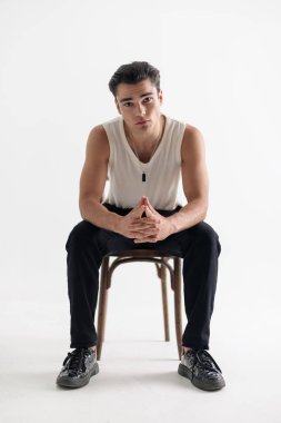 Dirseklerini dizlerinin üstüne çöküp oturan ve beyaz arka planda ileriye bakan genç sıradan bir adam.