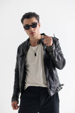 Siyah deri ceketli, gri arka planda dikilirken havalı bir şekilde poz veren, güneş gözlüklü kendine güvenen genç adam.