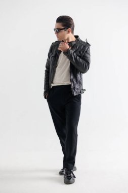 Güneş gözlüklü havalı genç adam yan tarafa bakıyor ve beyaz arka planda el ele yürüyor.