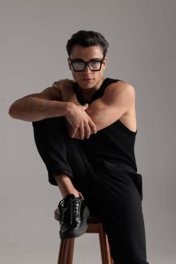 Gözlüklü, omuzlarına ve dirseğine dokunan ve dizleriyle gri arka planda oturan seksi kaslı adam.