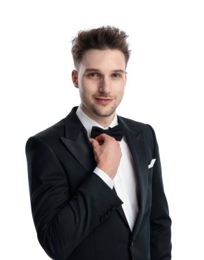 Smokinli mutlu genç damat ileriye bakıyor, siyah papyonu ayarlıyor ve beyaz arka planda gülümsüyor.