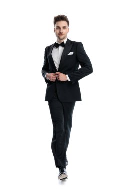 Önüne bakıp gülümseyen ve beyaz arkaplan önünde siyah smokini kapatan mutlu zarif damat.