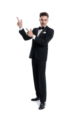Siyah smokinli kendine güvenen bir işadamı beyaz arka planda dururken parmaklarını havaya kaldırıp gülümsüyor.