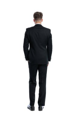 Siyah smokinli zarif genç damadın arka planda el ele tutuşup beyaz arka planın önünde duruşu.