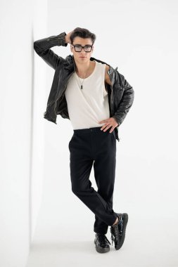 Siyah deri ceketli, gözlüklü, kafasının arkasında el ele tutuşan ve gri arka planda bir duvara yaslanan havalı moda adamı.