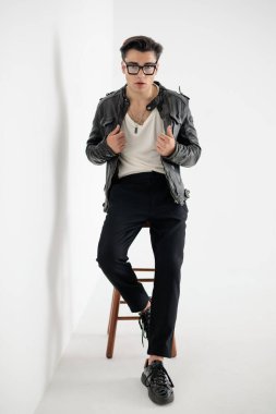 Gözlüklü, siyah deri ceketli ve gri arka planda oturan seksi havalı adam.