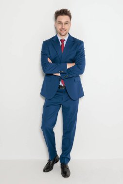 Mavi takım elbiseli mutlu, zarif bir adam gri stüdyo arka planında kollarını kavuşturmuş duruyor.