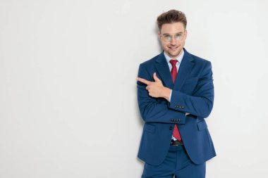 Mavi takım elbiseli, gülümseyen zarif genç adam, gri arka planda kollarını kavuşturarak parmağını yana doğru gösteriyor.