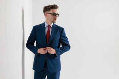 Güneş gözlüklü, ayakta duran, ceketini kapatan ve gri duvar arkasına bakan ciddi bir iş adamı..