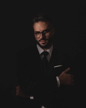 Takım elbiseli ve gözlüklü yakışıklı adam siyah stüdyo geçmişinde kollarını kavuşturarak poz veriyor.