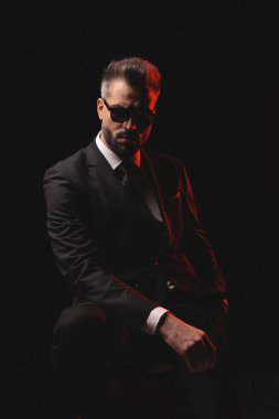 Güneş gözlüklü ve takım elbiseli ciddi bir adam kırmızı ışıkla arka planda el ele tutuşurken sandalyede poz veriyor.