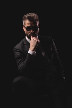 Güneş gözlüklü ve takım elbiseli dalgın bir adam oturuyor ve parmaklarını dudaklarında gezdirirken diğer elini koyu arka planda tutuyor.