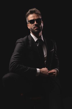 Kendine güvenen, güneş gözlüğü takan, sandalyede oturan ve siyah stüdyo arka planında takım elbisesini ayarlayan zarif bir adam..