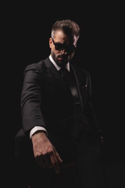 Takım elbiseli ve güneş gözlüklü şık bir adam siyah arka planda el ele tutuşurken sandalyede oturuyor.